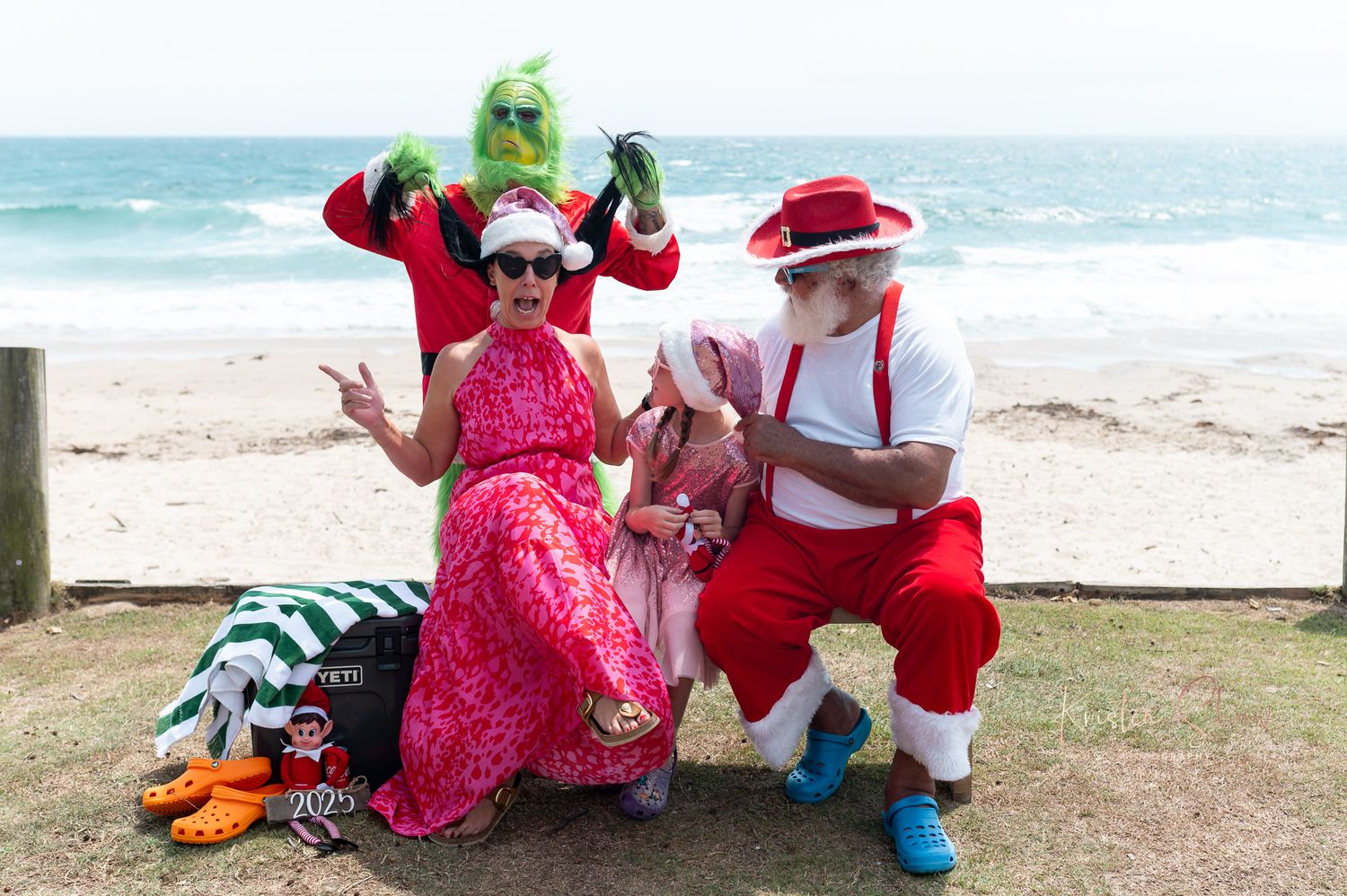 Colorful costumed performers in red and pink outfits pose playfully at a sunny beach location.