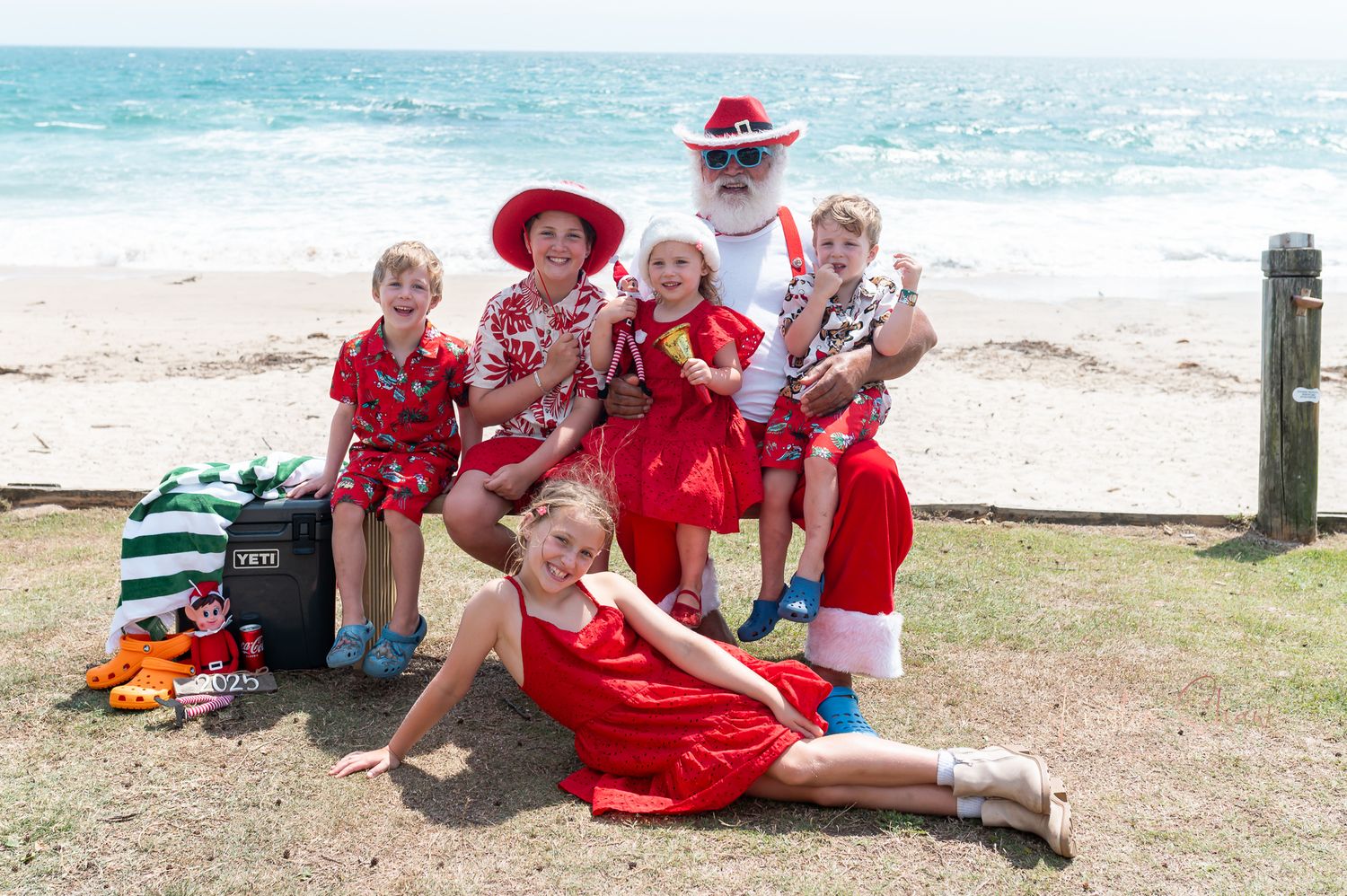 A group wearing red and white outfits poses together on grassy ground near a beach with ocean waves visible.