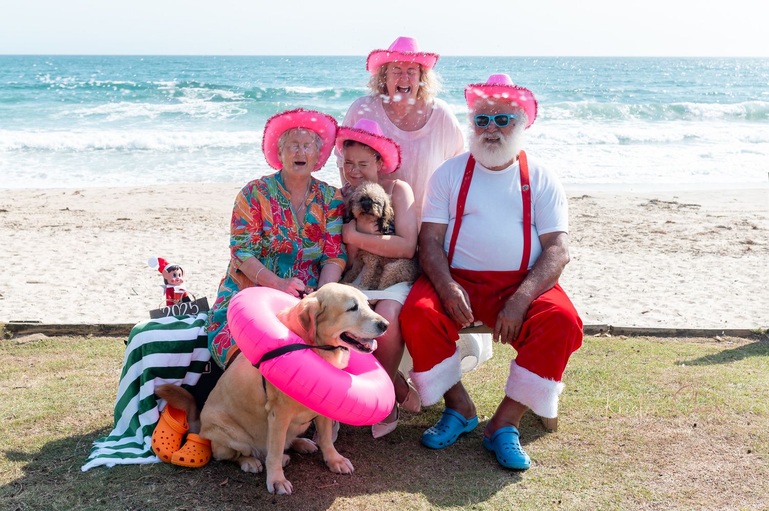 A group wearing red and white outfits sits with dogs on a sandy beach near the ocean.