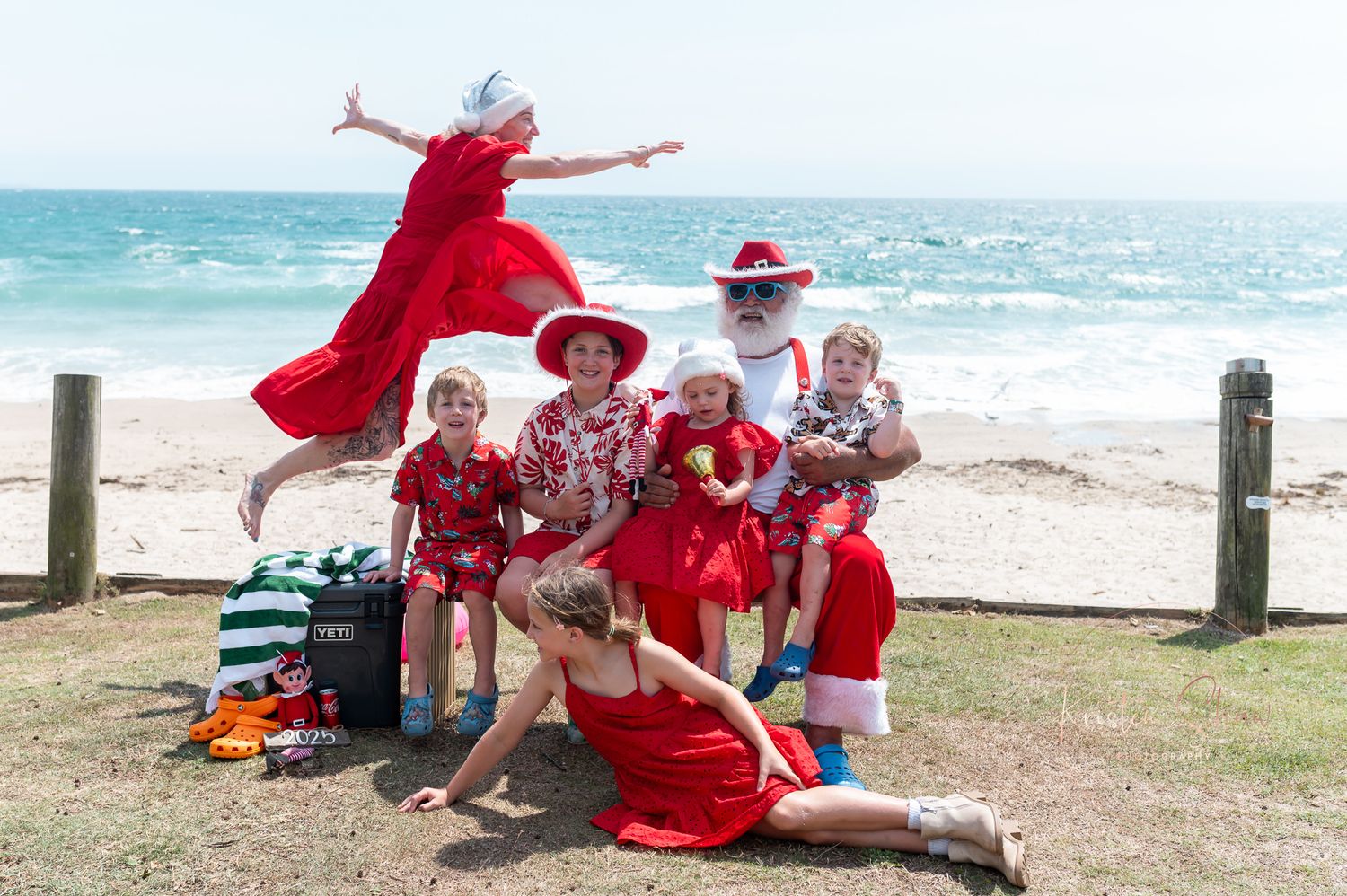 A group wearing red outfits gathers on a sunny beach near the ocean for a holiday celebration.