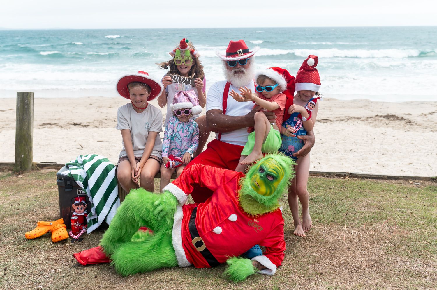 A group wearing festive Christmas attire and sun hats poses together on a grassy area by the beach.