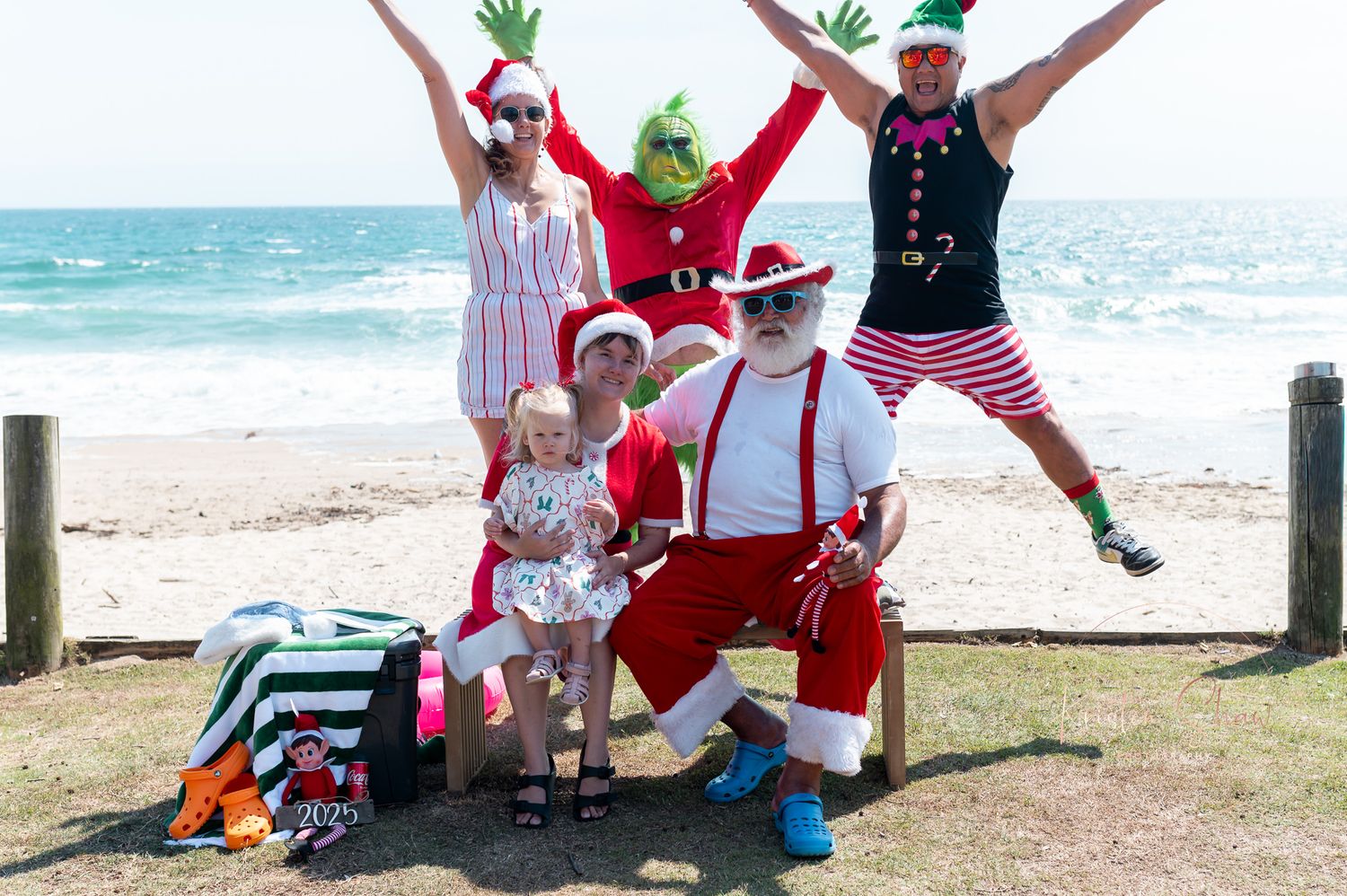 A group of people in holiday costumes jumping joyfully on a beach with ocean waves in the background.