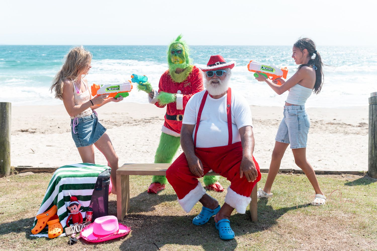 People gather on a sunny beach with Christmas decorations and Santa hats for a tropical holiday celebration.