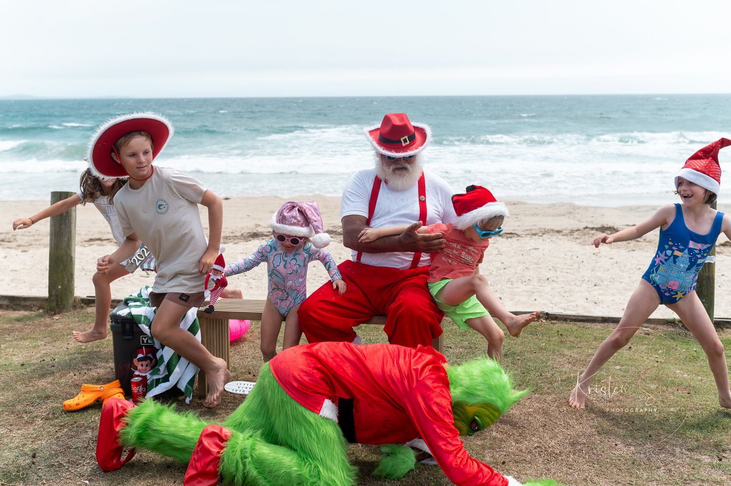 A group wearing Santa hats plays with green beach balls on a sandy beach near the ocean waves.