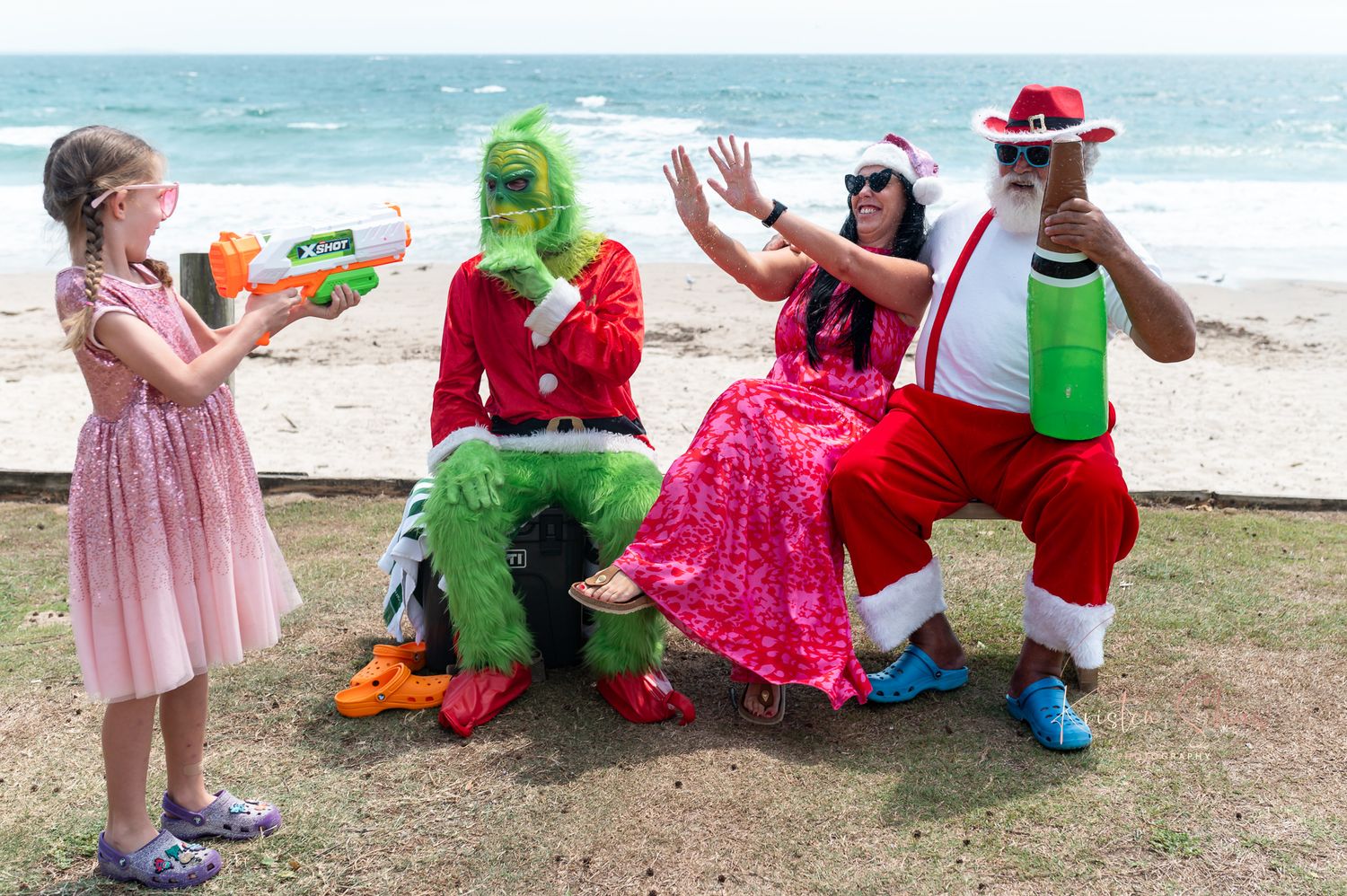 Whimsical beach photoshoot featuring holiday characters and pink accents against ocean backdrop.