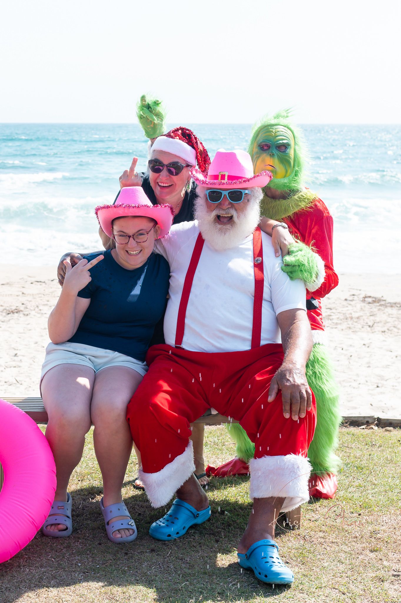 A festive beach scene with people in holiday costumes including red suspenders and Santa hats posing by the ocean.