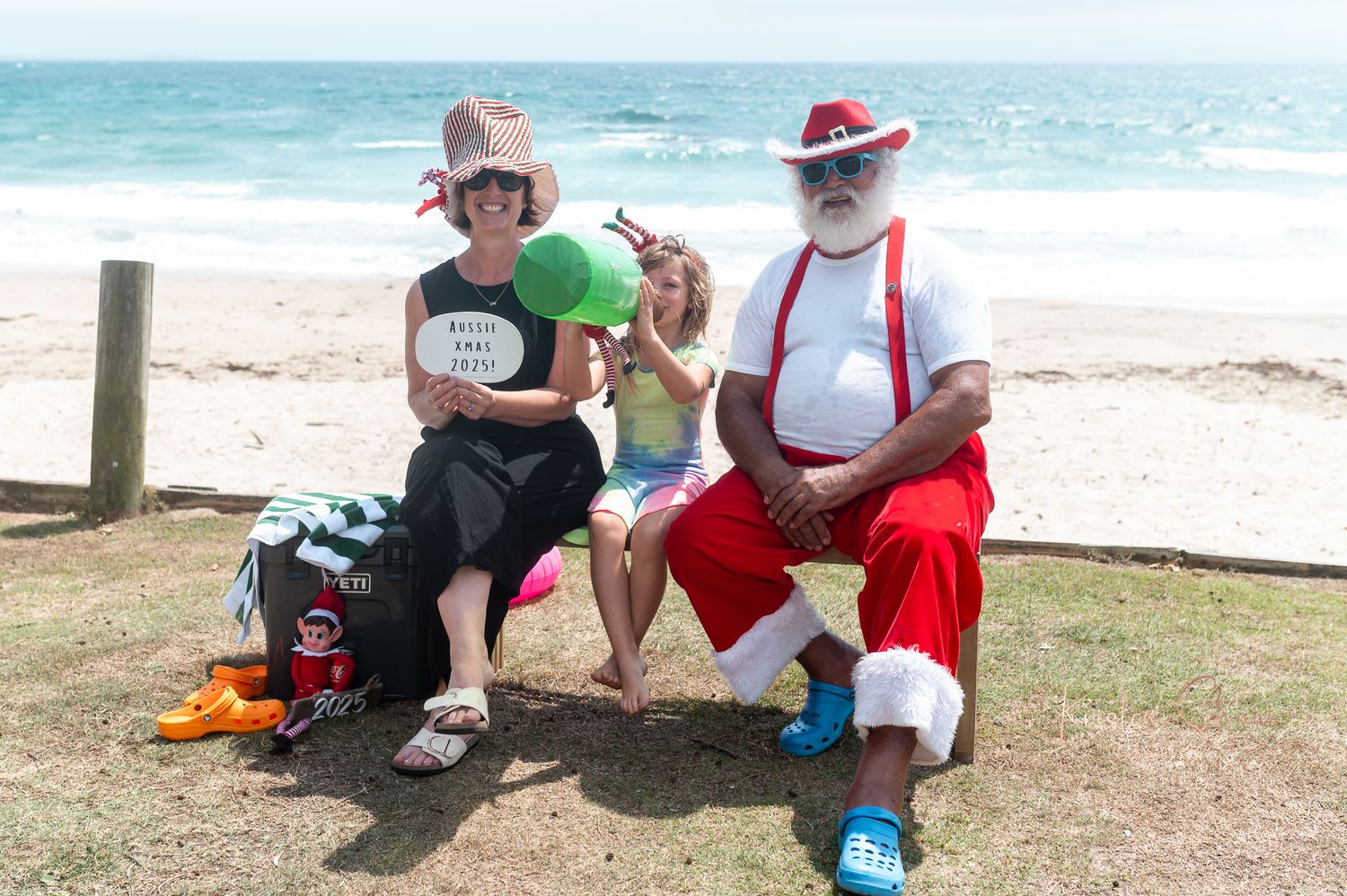 Beachgoers in festive summer outfits relax on the grass near the ocean with colorful accessories.