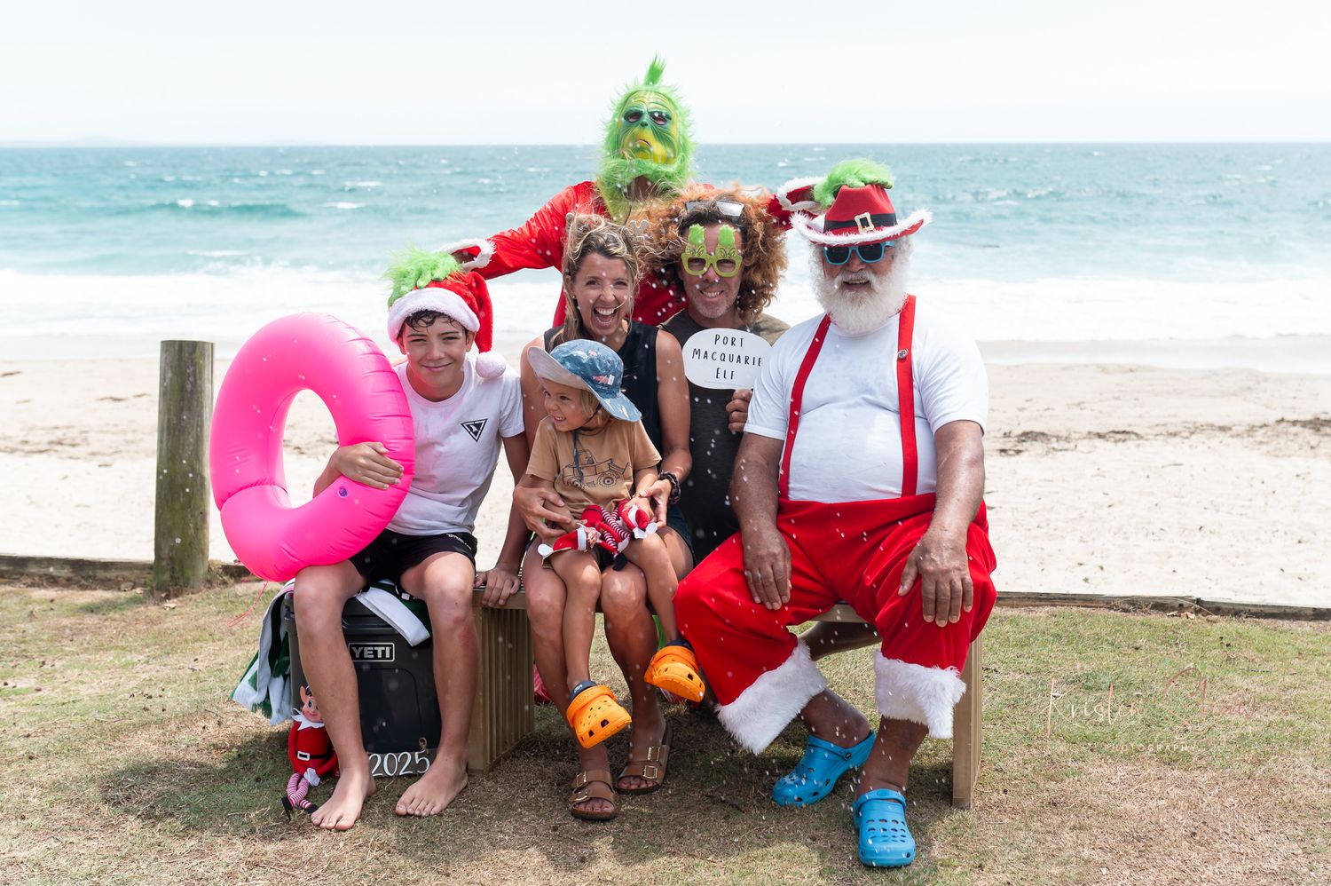 A cheerful group in holiday attire gathers on a beach with pink floaties and festive decorations.