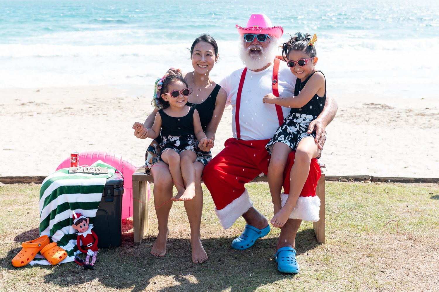 Group enjoying a beach day with bright summer attire including red shorts, suspenders, and a pink hat by the ocean.