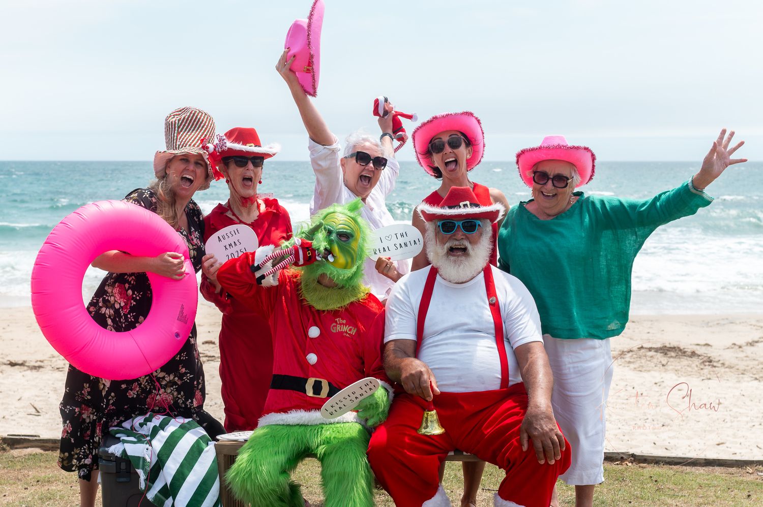 A group wearing festive holiday attire and colorful pool floats poses together on a sunny beach.