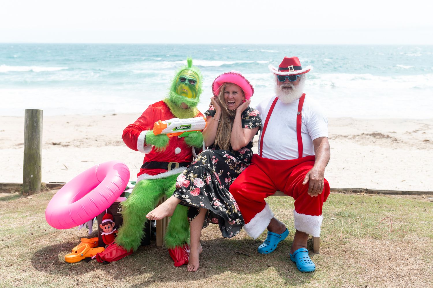 Festive characters in red and green costumes pose playfully on a sunny beach with ocean waves in background.