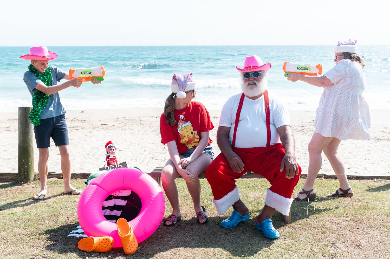 Group of people in festive attire with water guns and pink pool float having fun at sunny beach.
