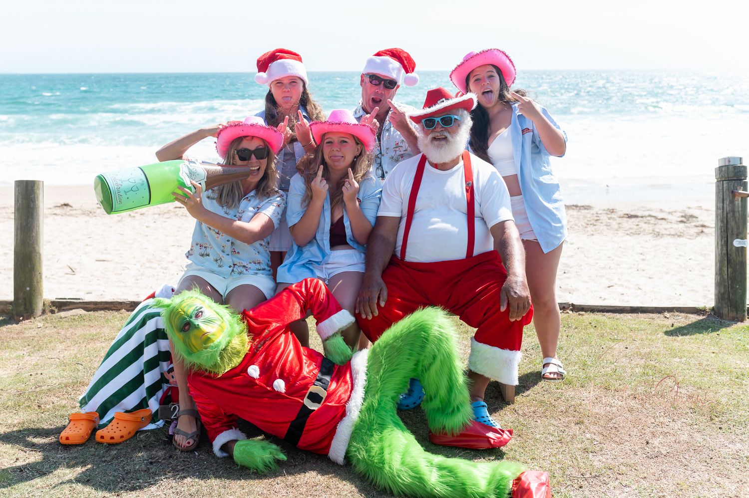 A group wearing red and white outfits sits on a beach while someone in a green costume lays on the ground.