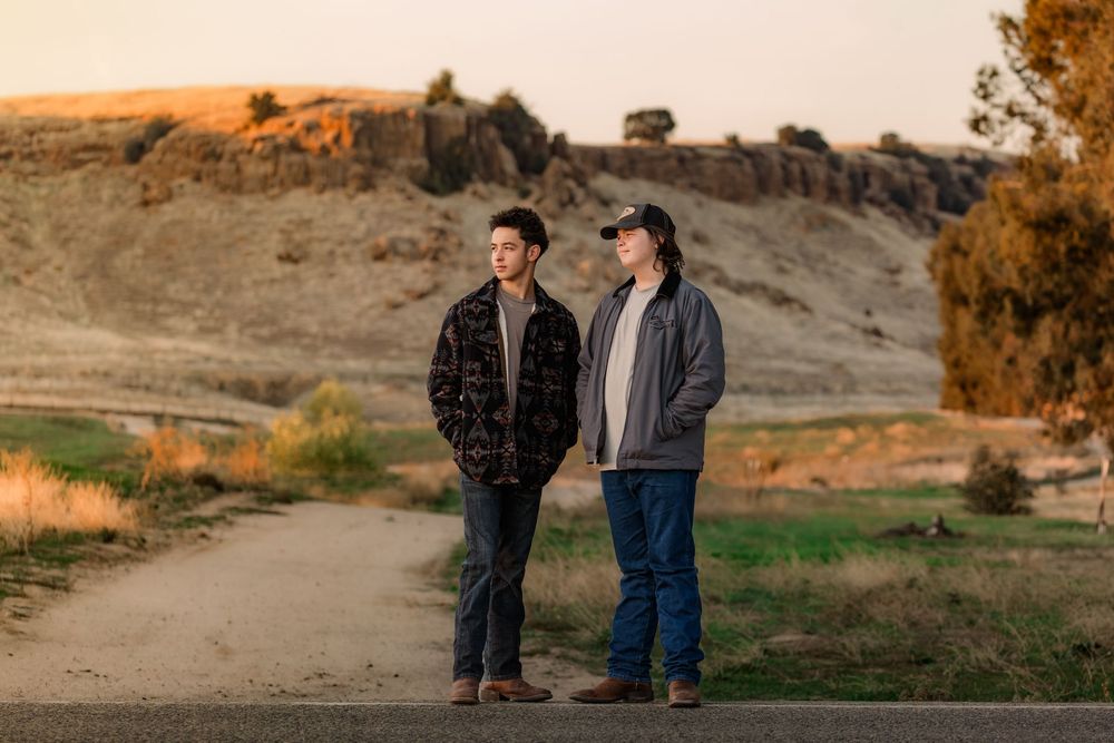 Two people stand together in a scenic desert landscape at sunset with mountains in the background.