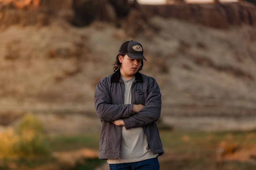Person in a dark baseball cap and jacket stands against a rocky outdoor backdrop in warm lighting.
