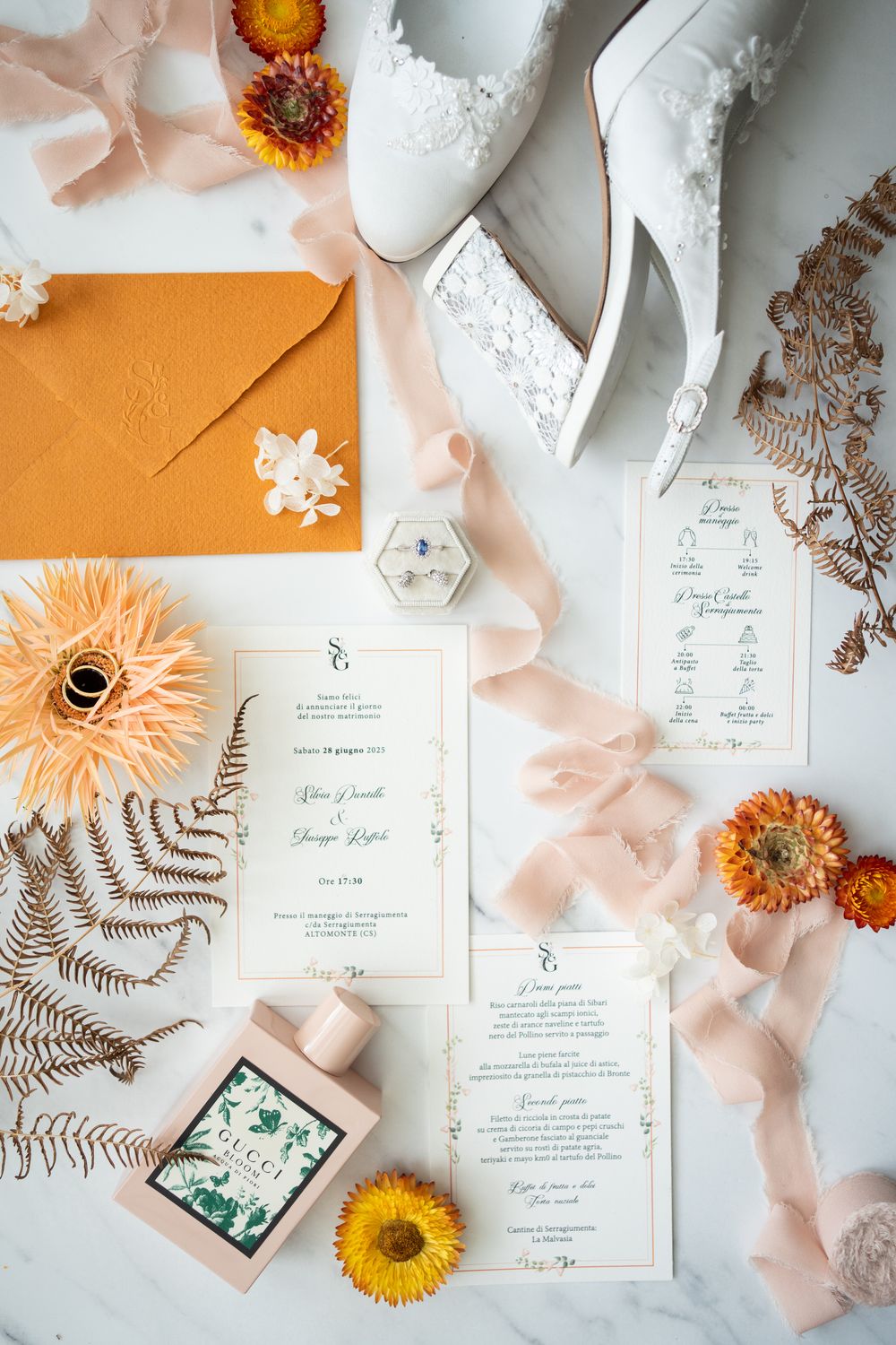 Wedding details flatlay with white lace shoes, orange flowers, invitation cards and peach ribbons on light background.