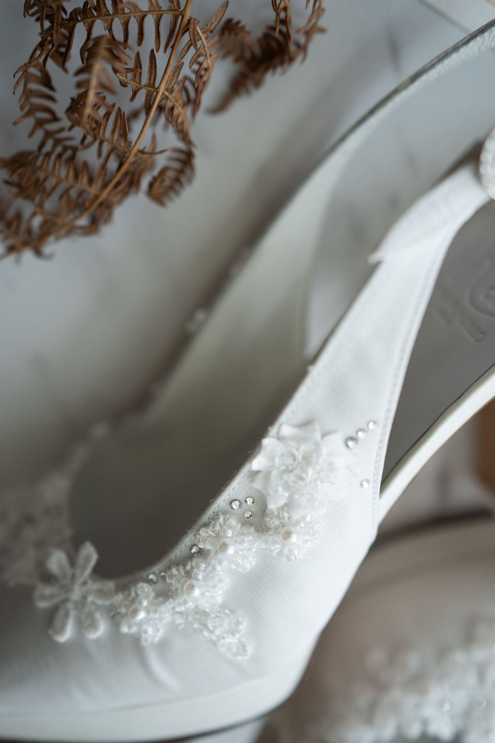 Elegant white bridal shoe with delicate lace embellishments and floral details displayed against a soft background.