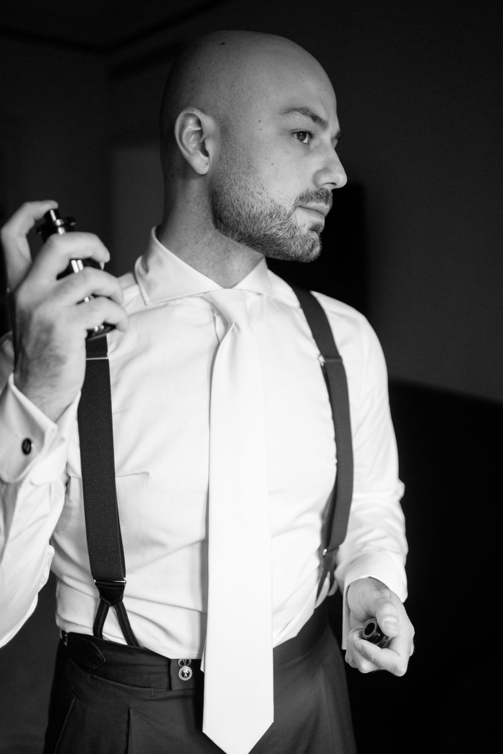 Black and white portrait of a person in formal attire with suspenders and tie applying cologne or perfume.