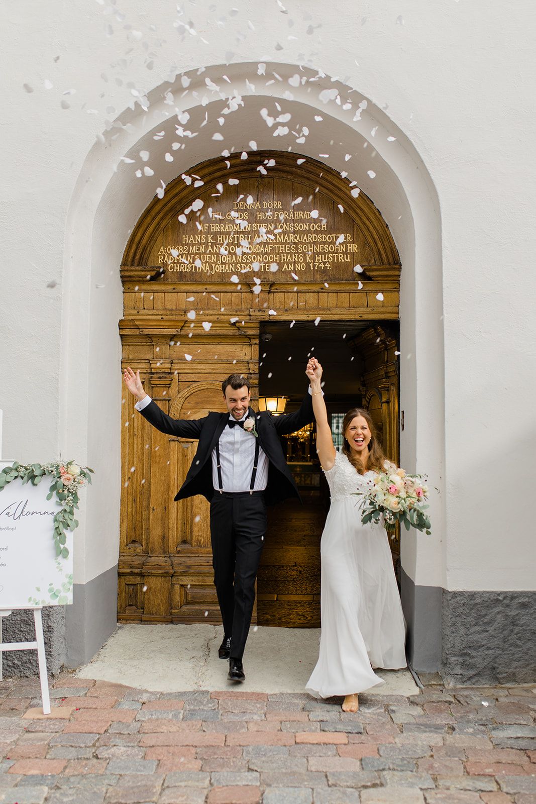 A couple celebrates joyfully under white stone archways while confetti falls around them during their wedding day celebration.