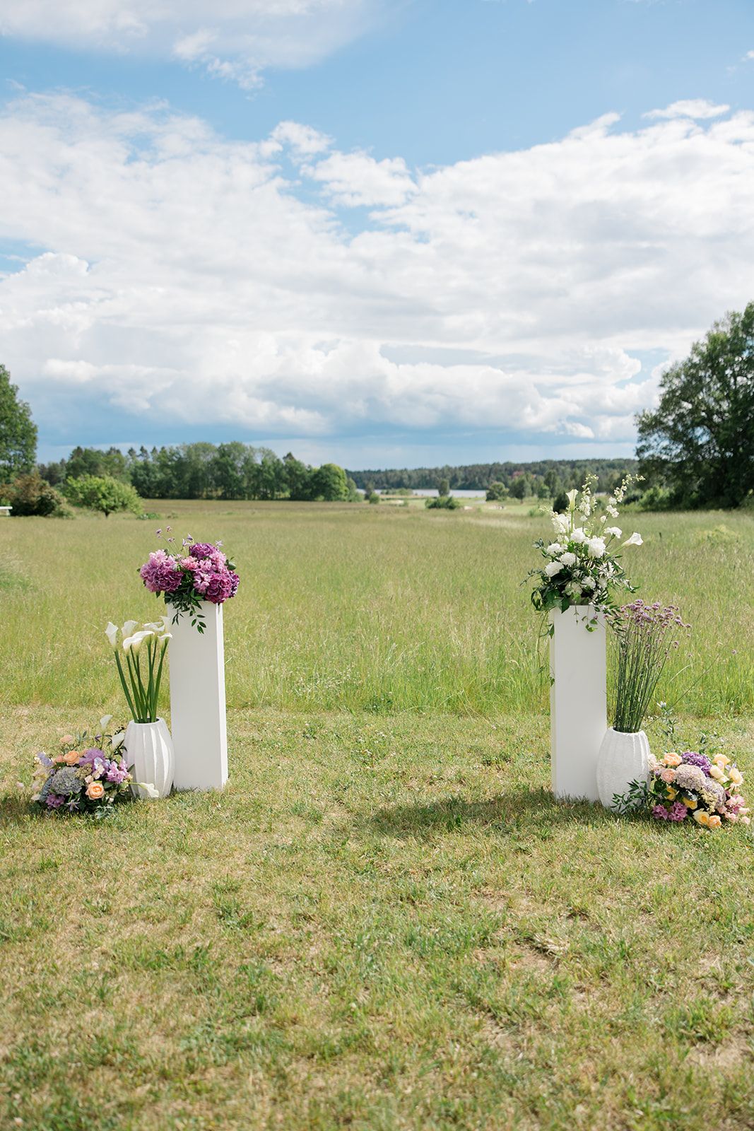 White ceramic vases with fresh floral arrangements stand in a serene meadow under blue summer skies.