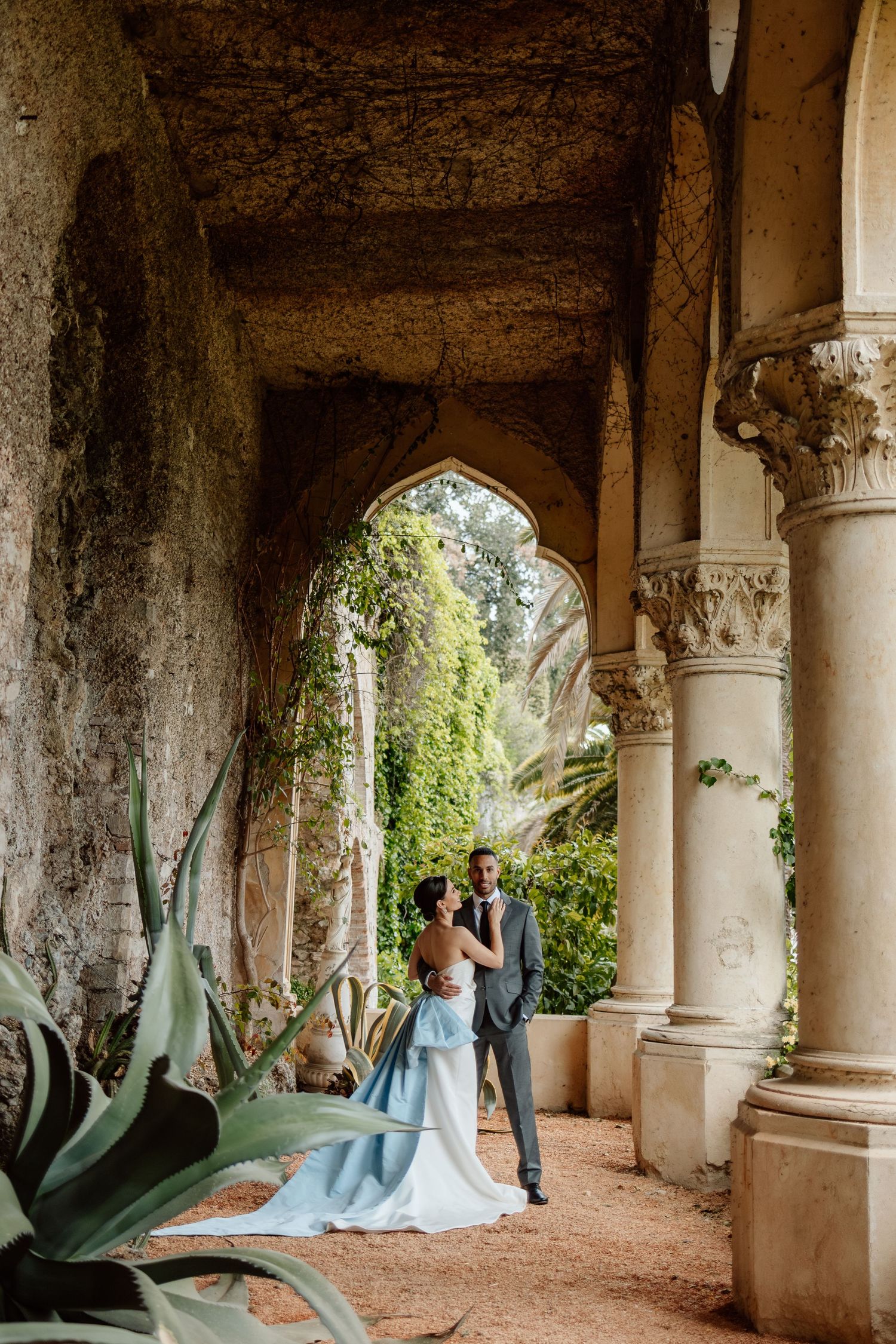 A couple stands under ornate stone archways in a lush garden setting with agave plants and vines at a romantic venue.