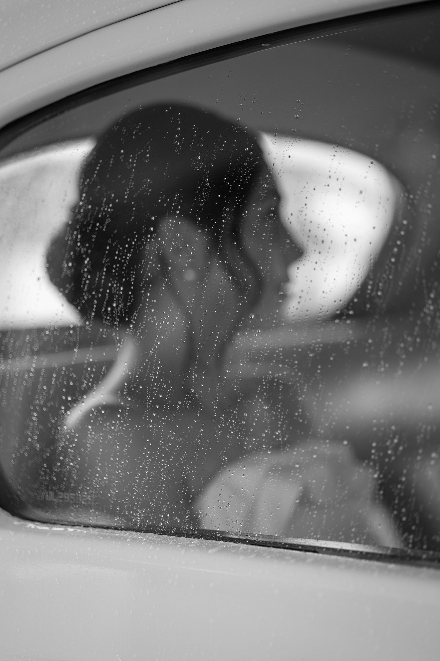 Black and white photo of a person sitting in a car viewed through a rain-covered window.