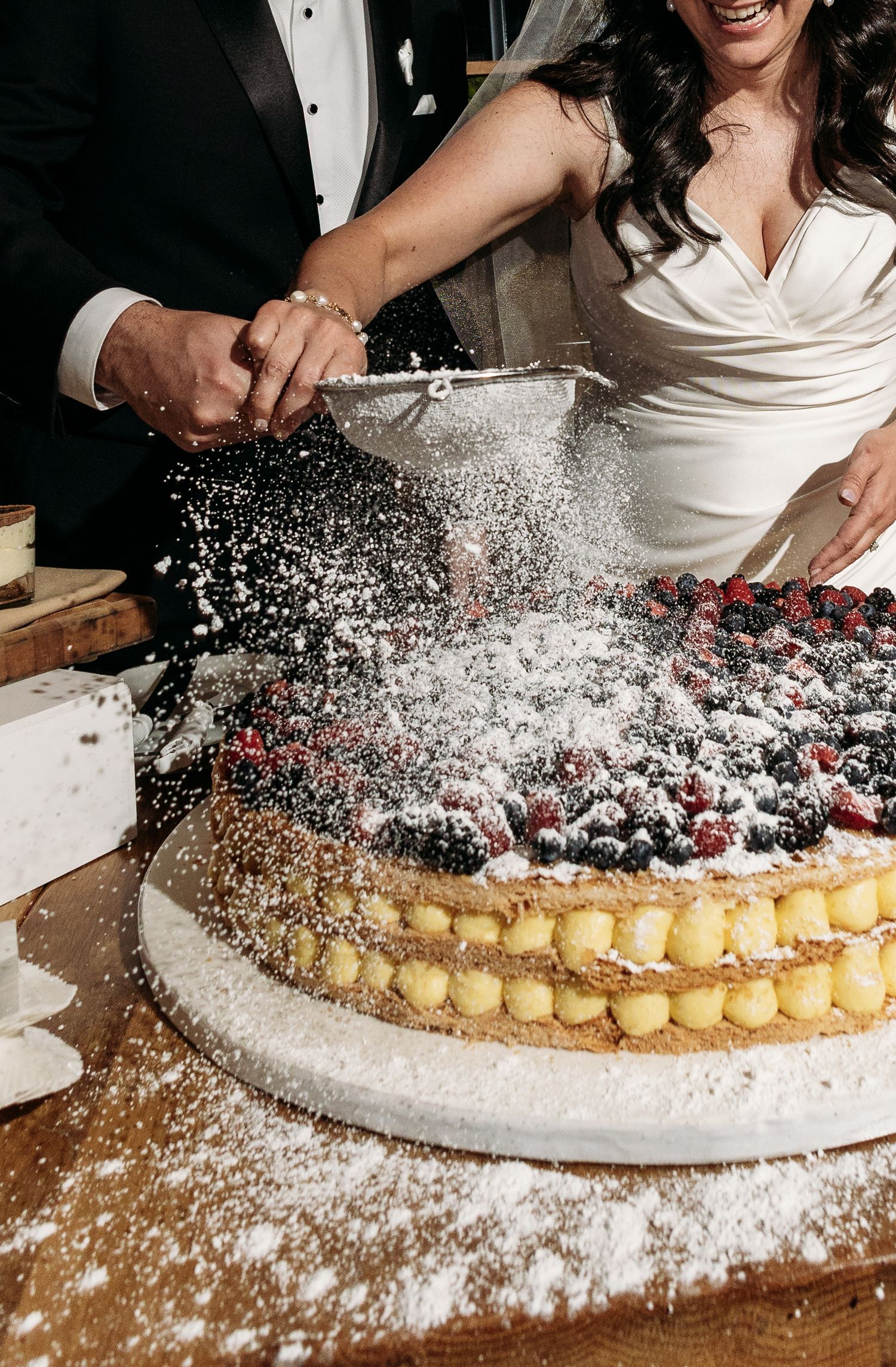 Someone dusts powdered sugar over a layered fruit tart at a wedding reception.