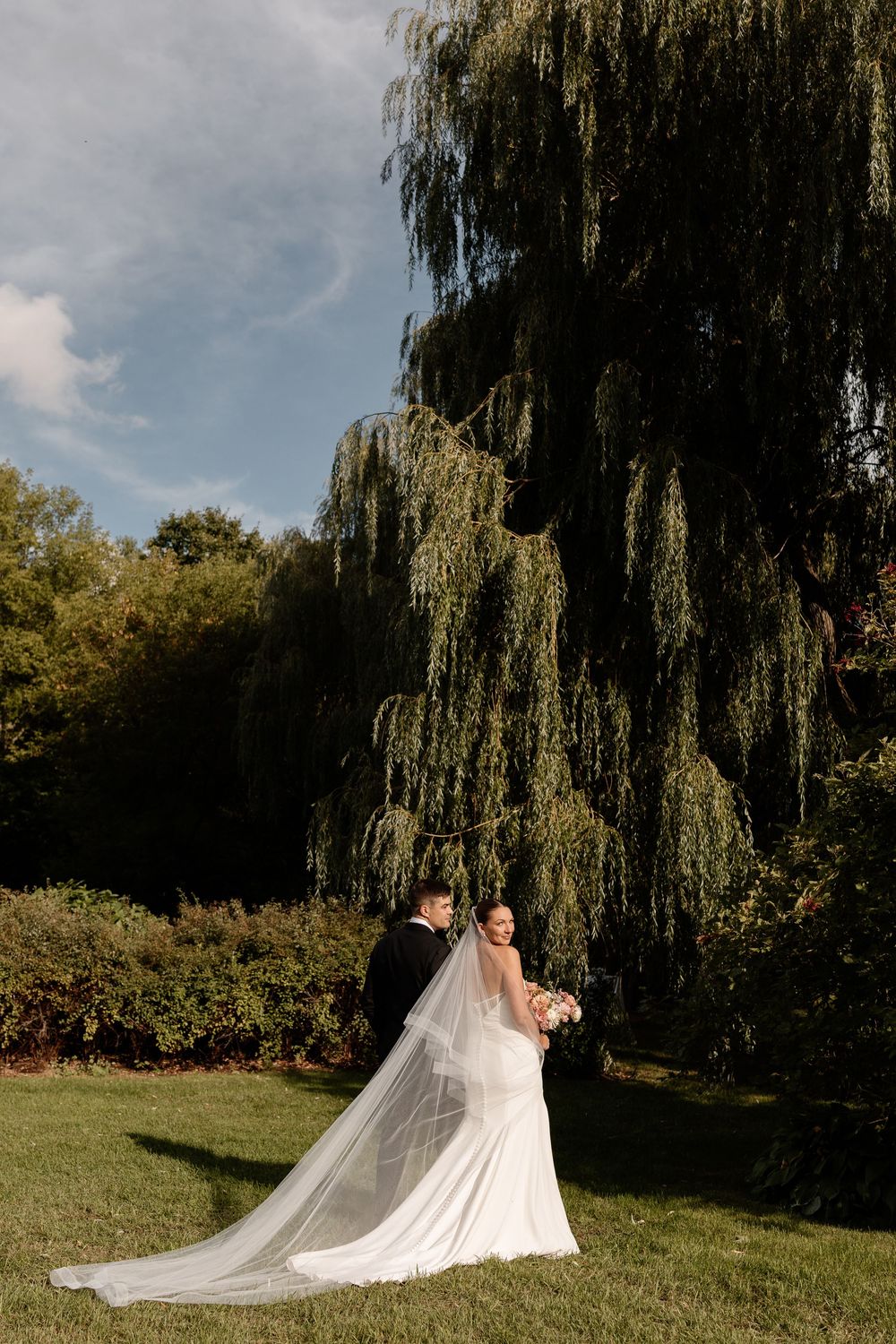 A bride in a flowing white gown stands beneath a graceful weeping willow tree in a sunlit garden setting.