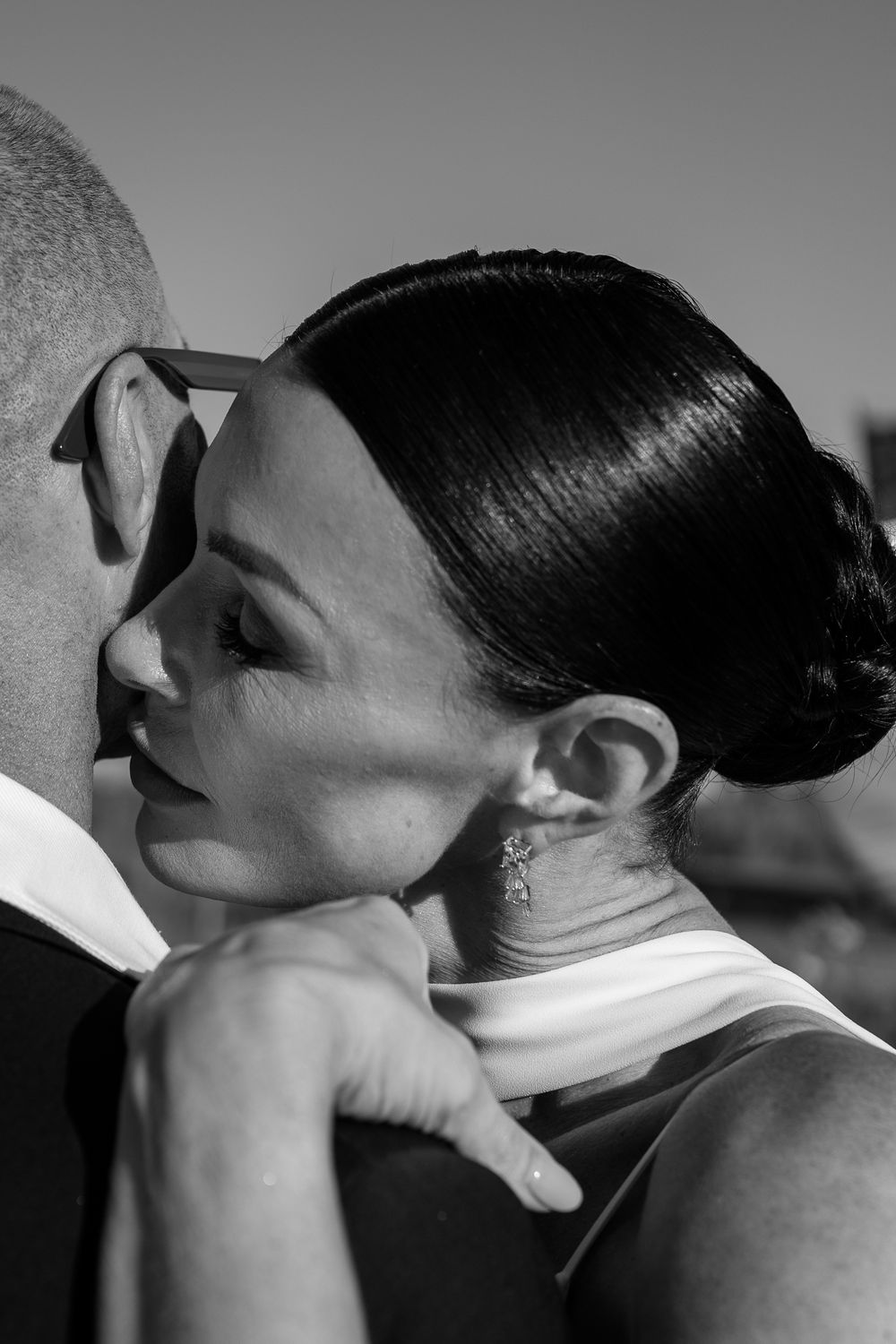Black and white photo of a bride resting her head on groom's shoulder, wearing elegant earrings at wedding.