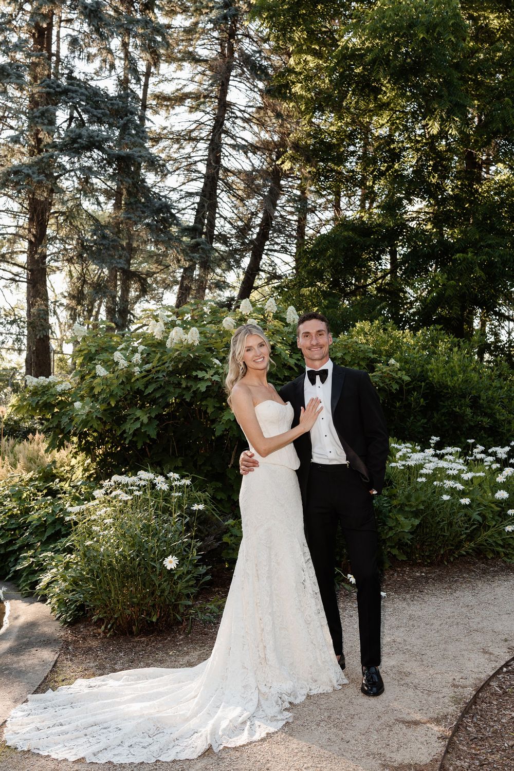 Bride in lace mermaid gown and groom in black tuxedo pose together in a lush garden setting.
