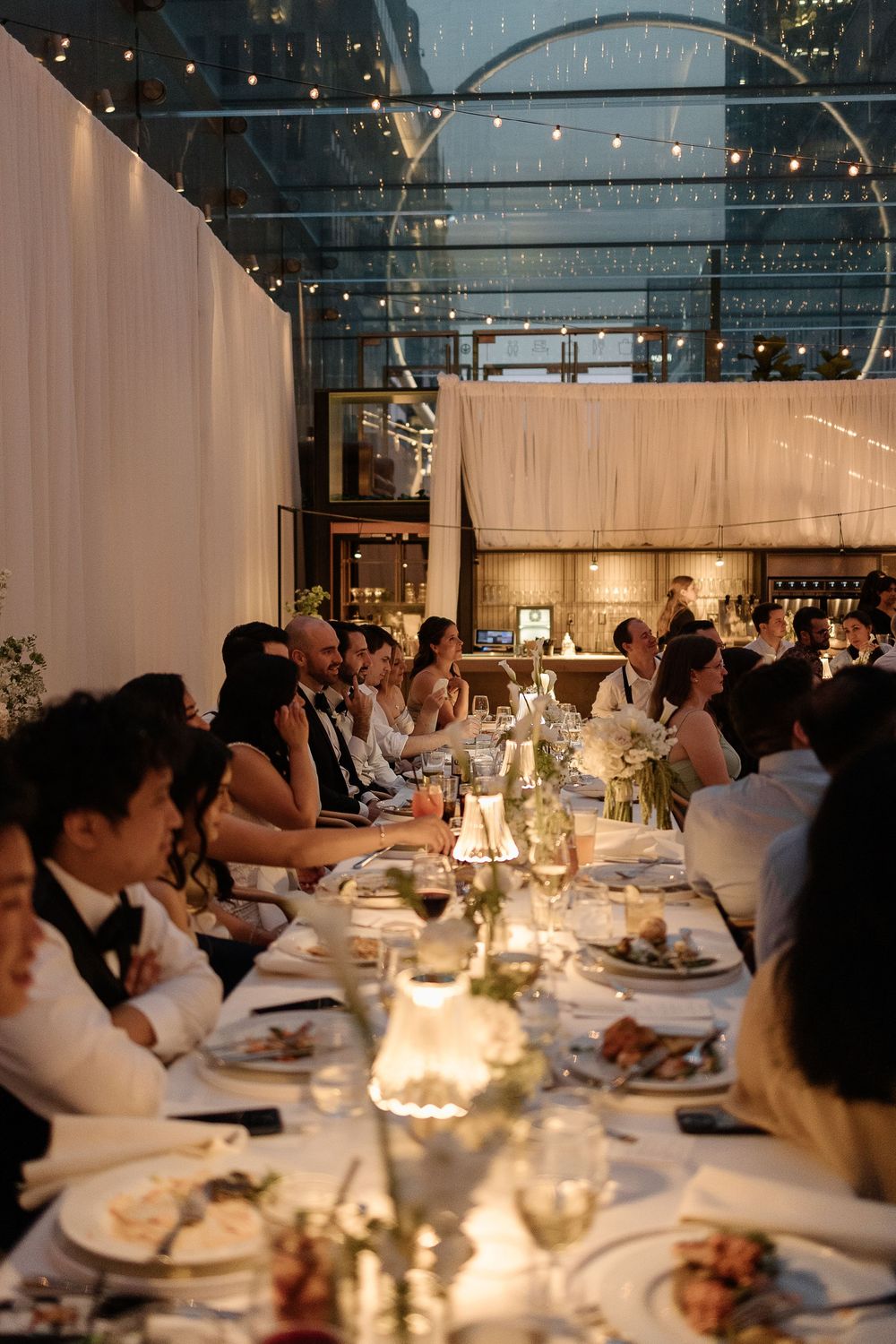Elegant candlelit dinner party with guests seated at a long white-clothed table in a glass-enclosed venue at night.