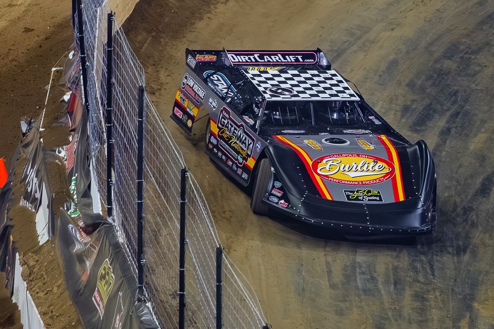 Race cars slide through a dirt track turn during a competitive stock car racing event on a muddy oval course.