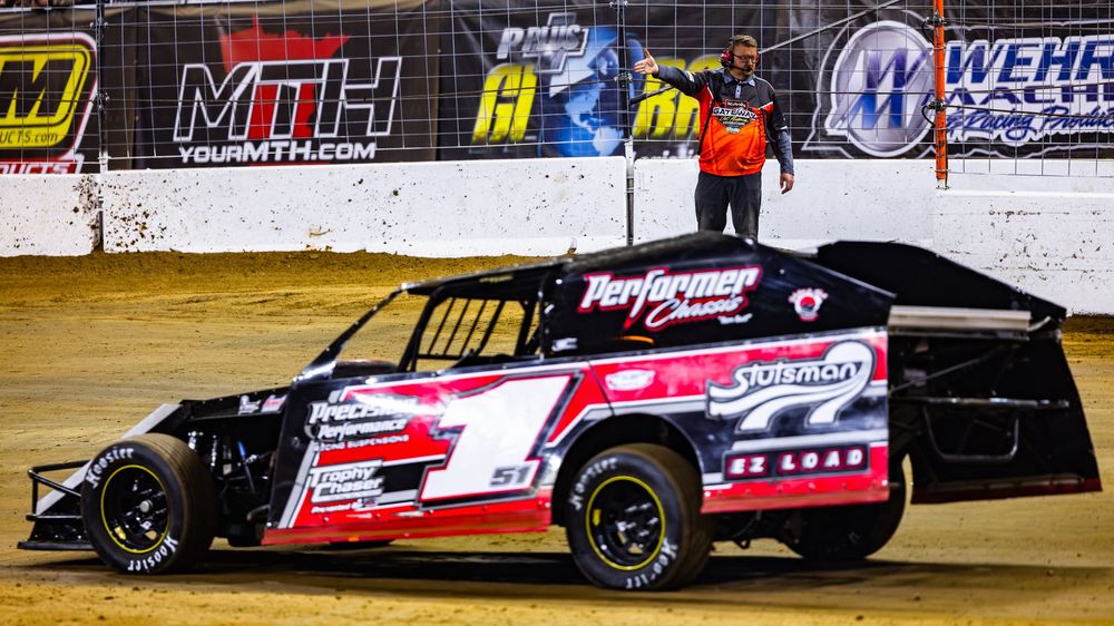Black and red modified dirt track race car speeds around corner on clay oval track with sponsor banners in background.