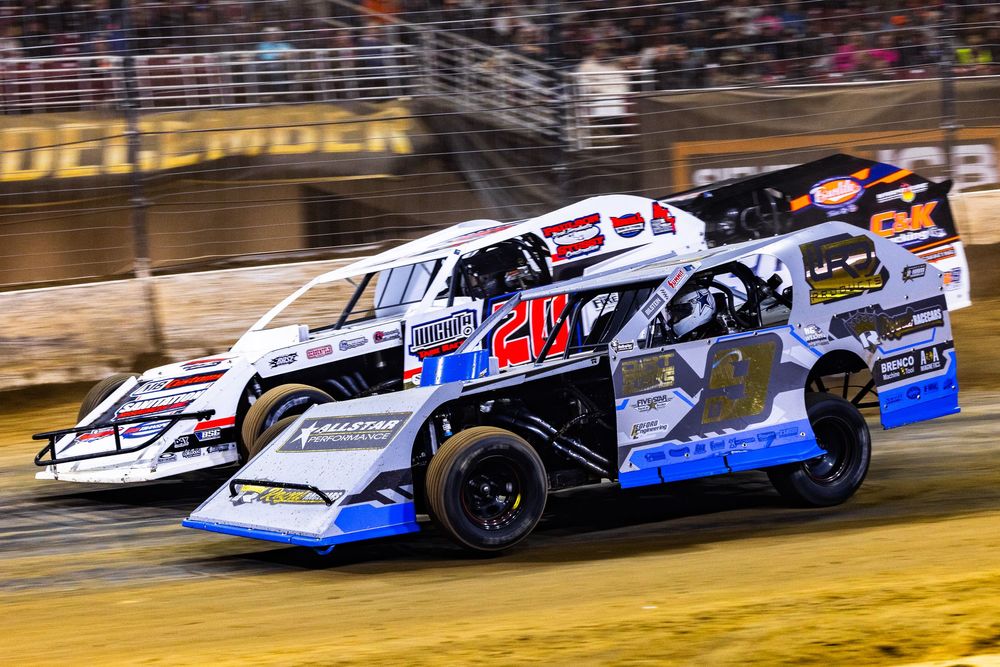Race cars speed around a dirt track during a competitive stock car racing event at night under bright lights.