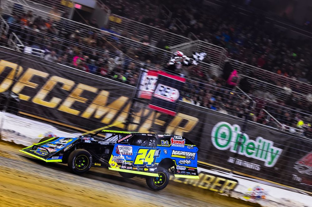NASCAR race cars speed around an oval track during a competitive night race under bright stadium lights.