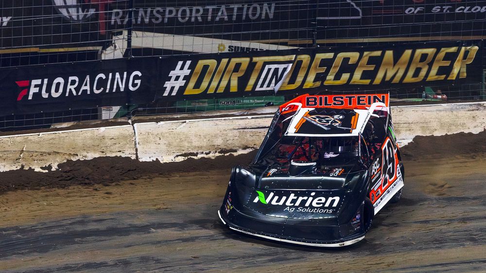 Dirt track race cars speed around a banked oval turn during a nighttime racing event.