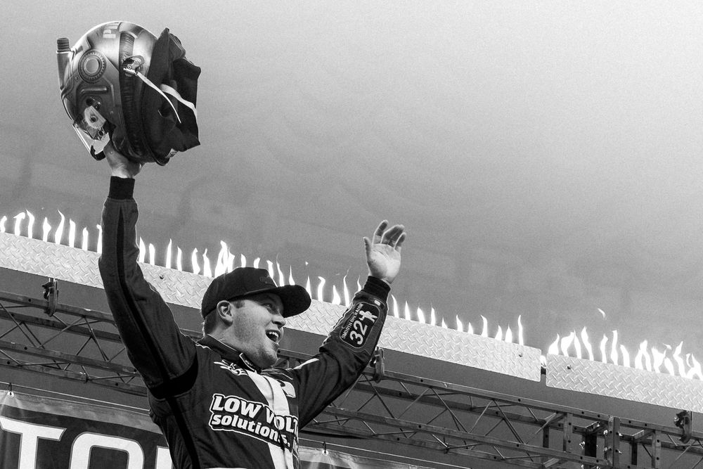 A race car driver celebrates victory with arms raised and helmet held high at the race track under dramatic skies.