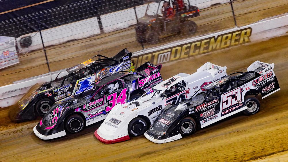 Late model dirt track race cars lined up in formation on a clay racing surface during a motorsports event.