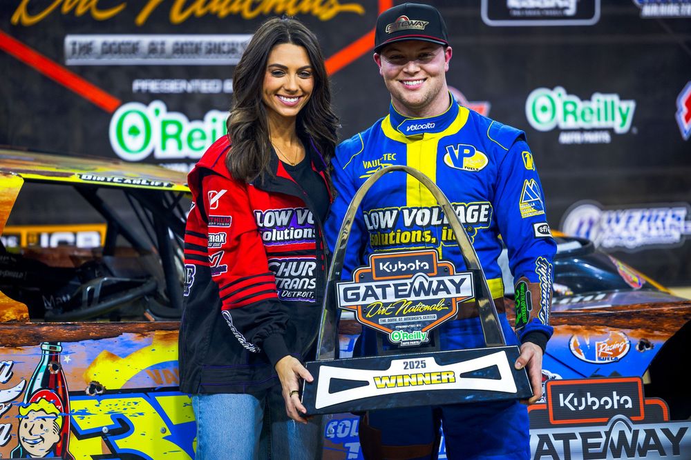 Two race car drivers pose with trophies and sponsor banners at a dirt track racing victory celebration.