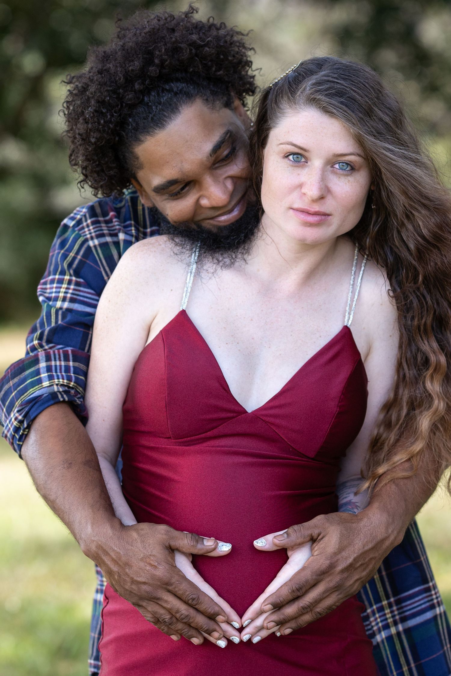 A couple embraces outdoors while wearing a burgundy dress and plaid shirt in a romantic maternity photo.