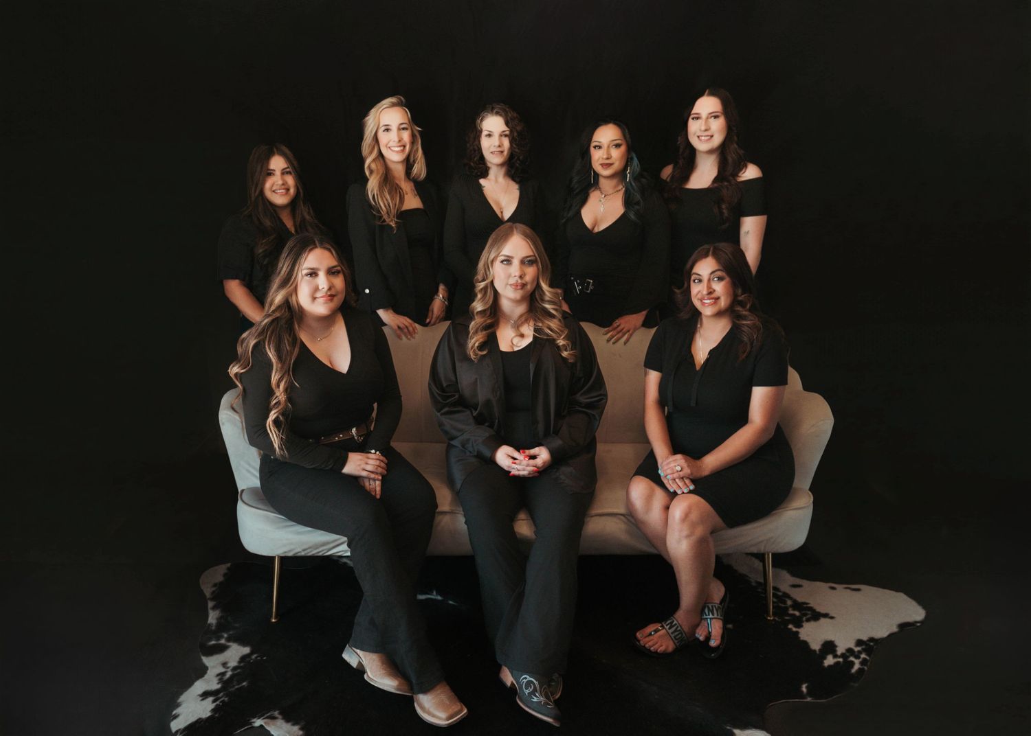 Eight women in matching black outfits pose together on a cream sofa against a dark backdrop, creating an elegant professional team portrait.