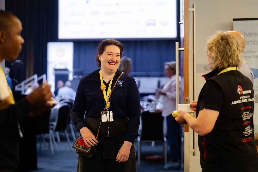 A person wearing a black t-shirt and name badge gestures while speaking to others in a conference, Fremantle Conference Photography