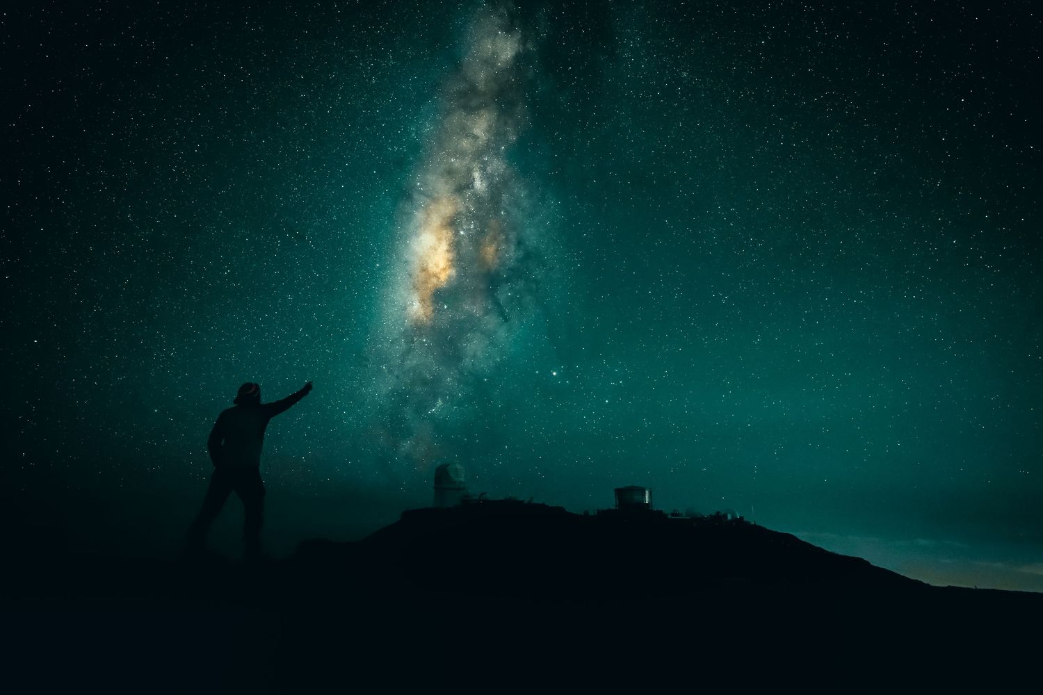 Silhouetted figure stands atop a hill against a stunning starry night sky with the Milky Way galaxy visible.