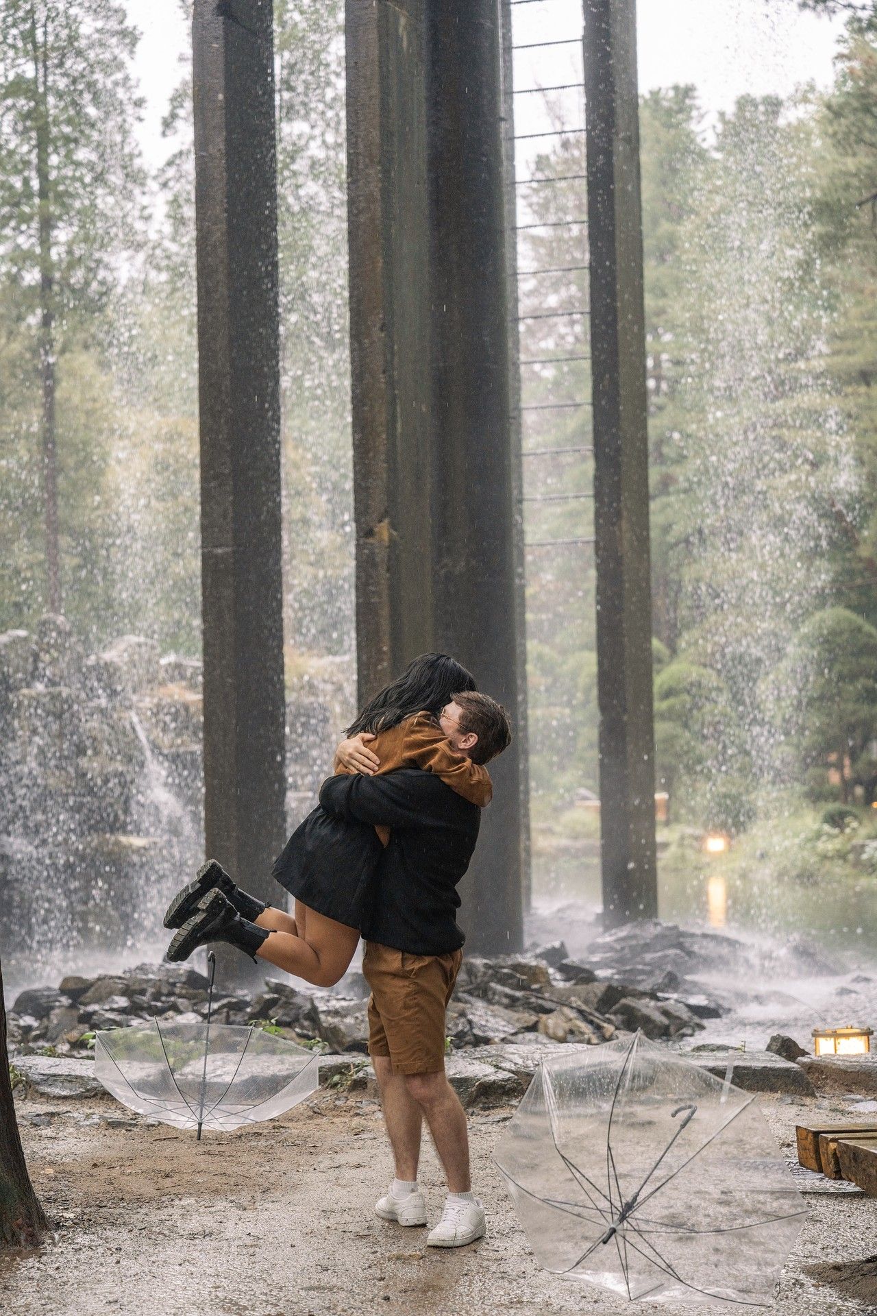 A couple embraces in a romantic moment among tall trees and misty spray in a forest setting.