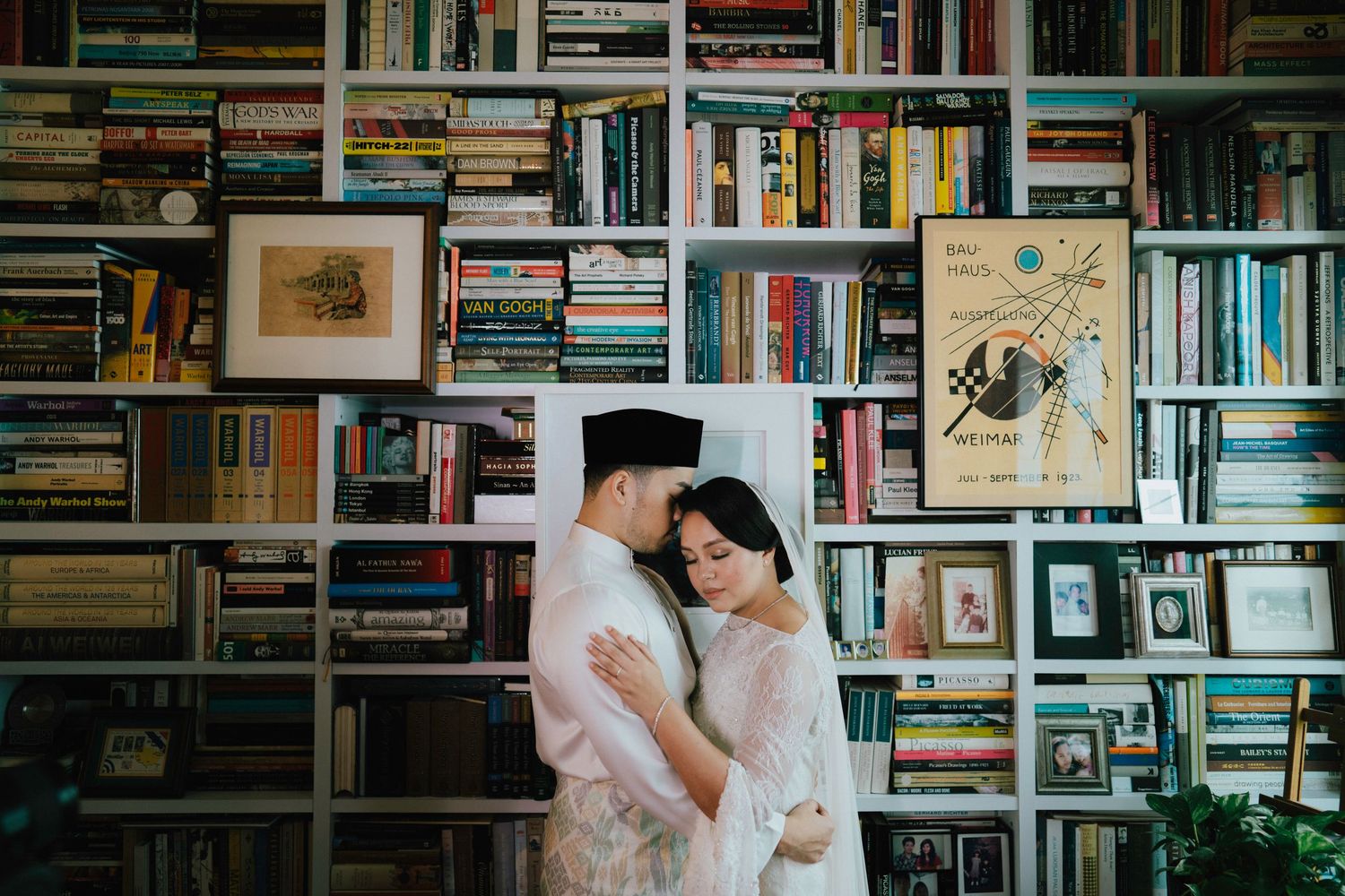 Couple in traditional Muslim wedding attire embracing tenderly in front of a large bookshelf wall.