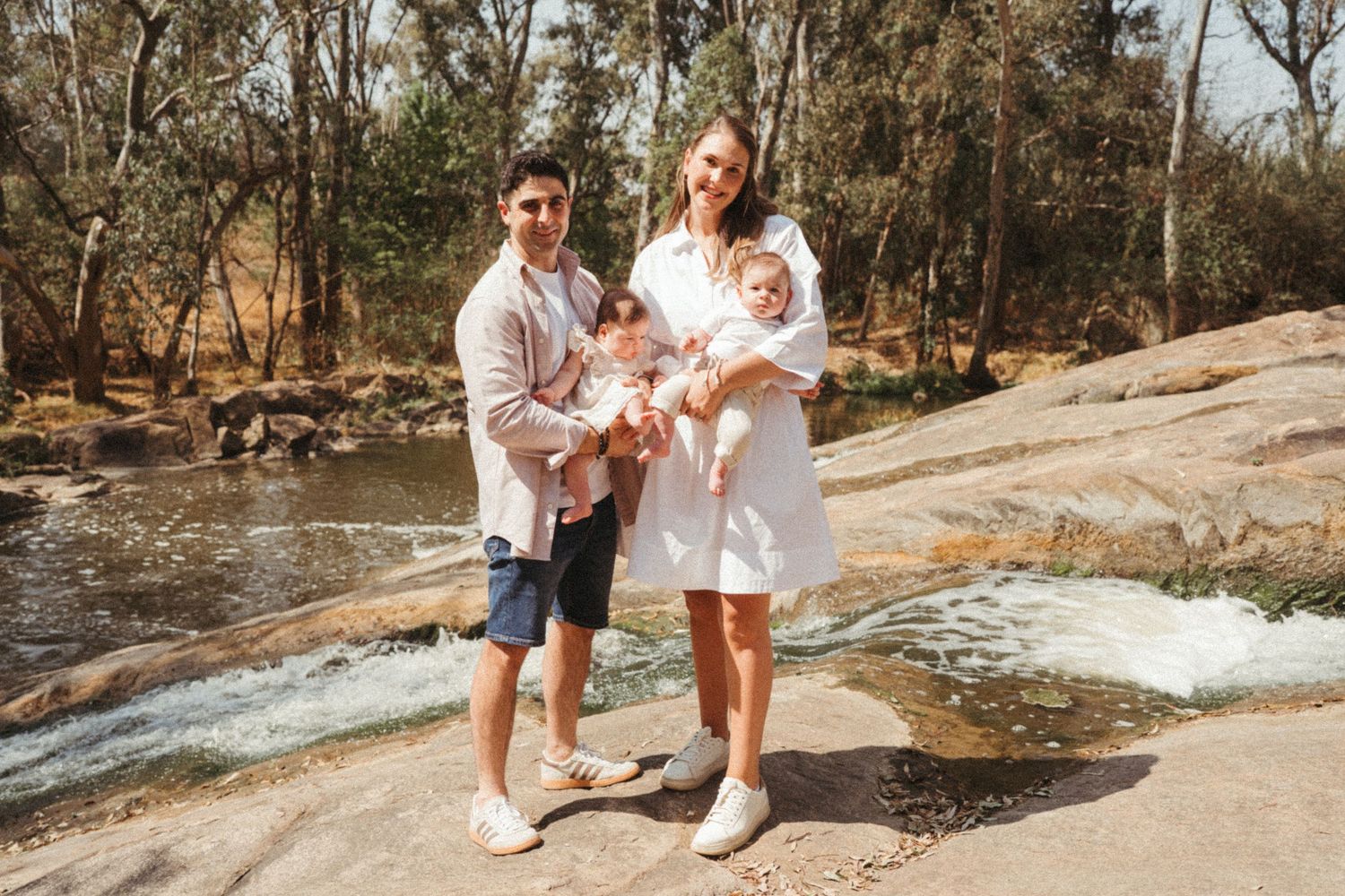 A family in white outfits poses together on rocky terrain near a stream in a natural outdoor setting.