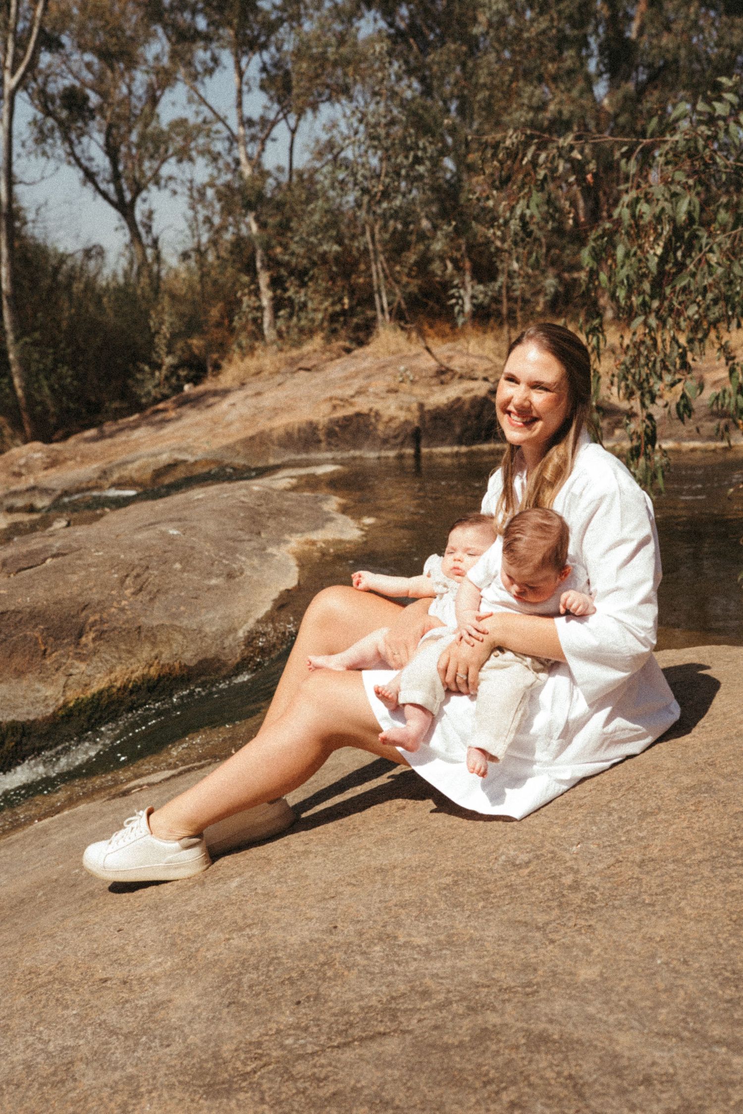 A parent and baby in white clothing sit together on a rocky outcrop by a stream on a sunny day.