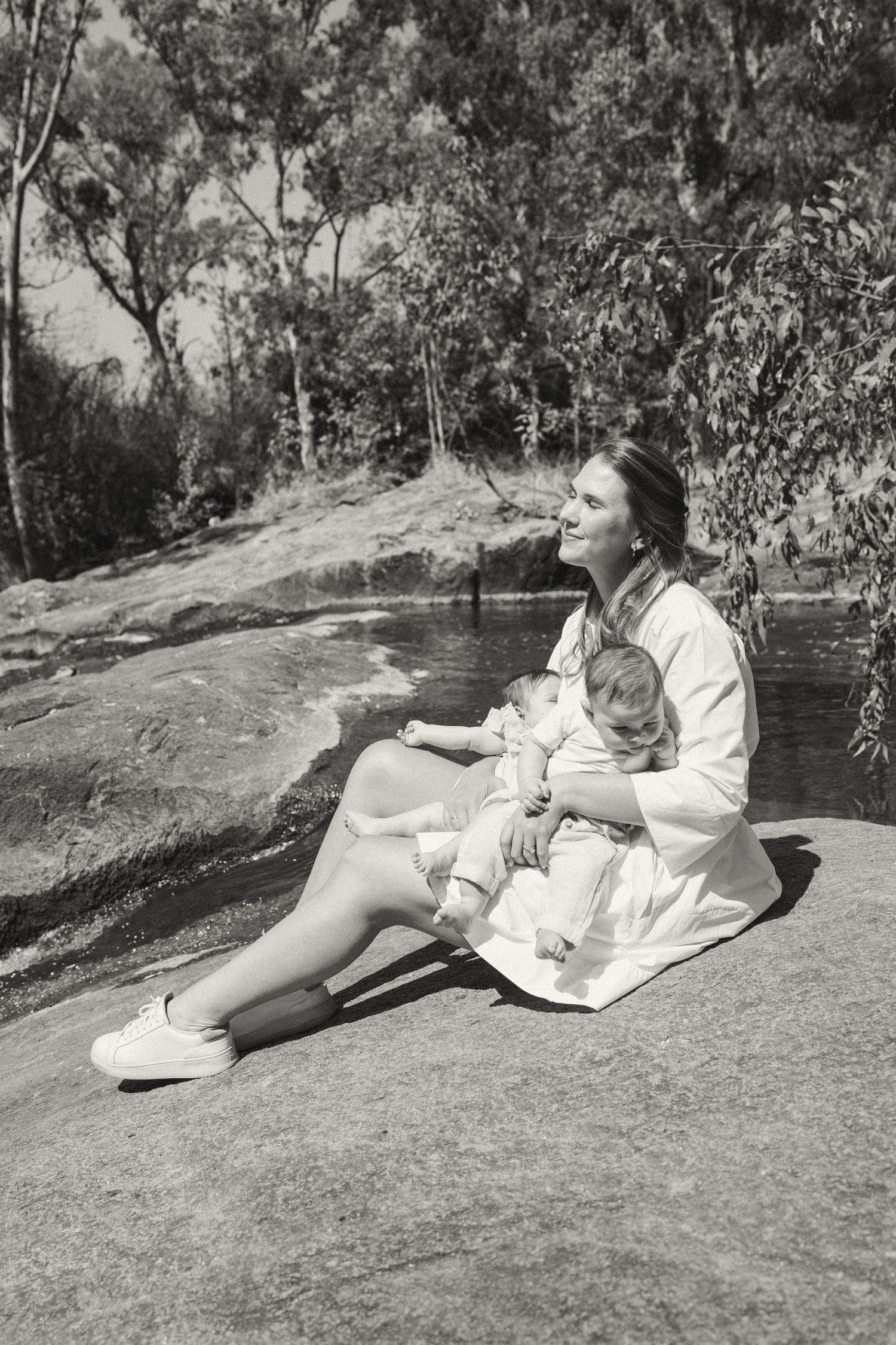 A black and white photo shows a parent and infant relaxing on a rocky surface near water.