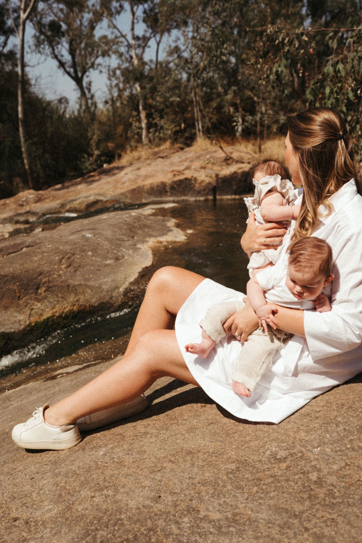 A parent in a white dress holds a baby while sitting on a rocky surface near water in nature.