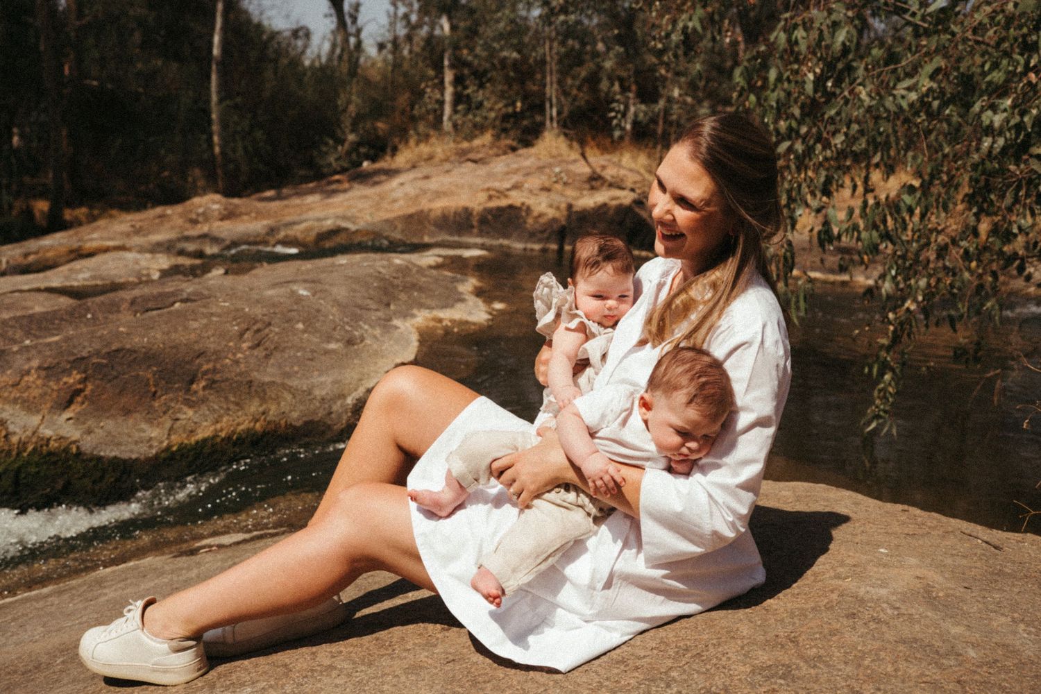 A parent in white clothing sits with two babies on a rocky surface near water in a wooded setting.